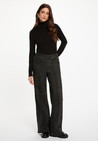 Schwarzer Rollkragenpullover kombiniert mit weiten, gemusterten, dunkelgrauen Hosen. Die Hosen verfügen über komplizierte Details und Taschen, ergänzt durch glänzende schwarze Schuhe.