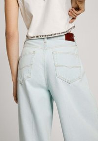 Valaistut leveälahkeiset farkut, joissa on teksturoitu kangas. Takana on kaksi taskua ja hienovaraiset ompelukohdat. Vyötäröllä ruskea nahkainen merkki.