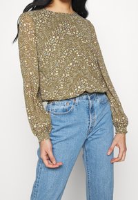Olivgrüne Bluse mit floralem Muster, langen puffigen Ärmeln und rundem Ausschnitt, kombiniert mit hellblauen Jeans.