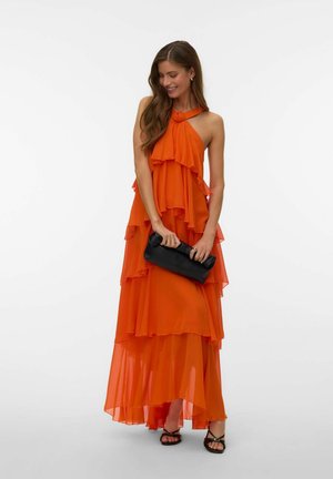 Femme portant une robe orange à volants nouée autour du cou, tenant une pochette noire, chaussée de talons noirs à lanières, souriante et regardant vers le bas.