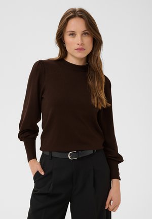 Vrouw met lang lichtbruin haar die een donkerbruin shirt met pofmouwen en een zwarte broek met riem draagt, staand tegen een effen achtergrond.