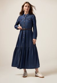 Abito in denim con colletto, chiusura a bottoni frontale e due tasche sul petto. Presenta una gonna a balze e una cintura marrone. Indossato con sneakers chiare.