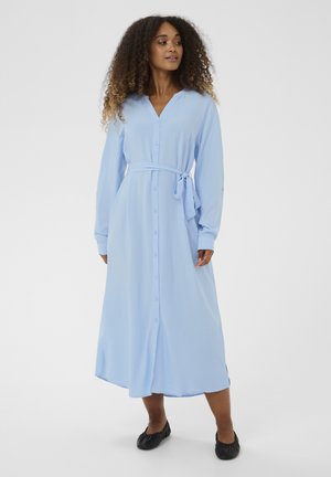 Ženska s kodrastimi lasmi, oblečena v dolgo svetlomodro obleko z gumbi in pasom v pasu ter črnimi ravnimi čevlji, stoji na belem ozadju.