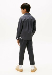 Camisa de botón gris oscuro con mangas largas, detalle en el yugo trasero, combinada con pantalones a juego y zapatillas beige sobre un fondo blanco.