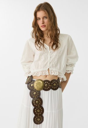 Ceinture en cuir marron avec des accents circulaires en or, une quincaillerie texturée, portée sur une jupe blanche fluide et une blouse blanche brodée.