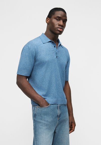 Νεαρός άνδρας με σκουρόχρωμο δέρμα που φορεί ανοιχτό μπλε κοντομάνικο πόλο πουκάμισο και ανοιχτό μπλε τζιν, στέκεται με το ένα χέρι στην τσέπη.