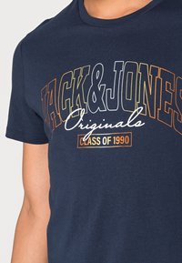Námorně modré tričko s krátkým rukávem, na přední straně s grafikou v oranžovém, bílém a žlutém textu: "JACK & JONES Originals TŘÍDA 1990."