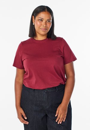 Femme aux cheveux noirs mi-longs portant des boucles d'oreilles argentées, un t-shirt bordeaux à manches courtes et un pantalon foncé taille haute, debout.