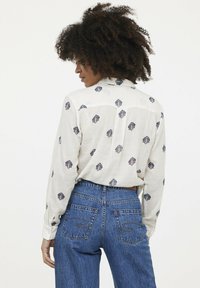 Chemise à manches longues en tissu crème léger, avec des motifs géométriques multicolores et un col classique, associée à un jean en denim taille haute.