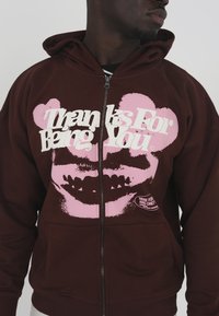 Brauner Reißverschluss-Hoodie mit pinken Text und einem grafischen Design, das ein Bärengesicht zeigt. Enthält Fronttaschen und ein Logo-Patch in der unteren rechten Ecke.