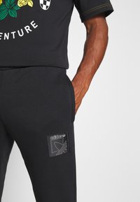 Černé sportovní kalhoty s hladkým povrchem, které mají na nášivce velké vyšívané logo Adidas, boční kapsy a přiléhavý střih.