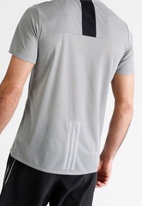 Camiseta deportiva de manga corta en gris claro con un panel negro en la parte trasera, que presenta tres rayas verticales blancas en el dobladillo inferior.