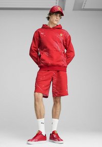 Haut à capuche rouge et short avec un design éclaboussé blanc, arborant un logo Ferrari. Porté avec des baskets rouges et des chaussettes blanches. Matériau en coton.