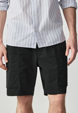 Mann trägt schwarze Cargoshorts und ein helles, langärmeliges Hemd mit hochgekrempelten Ärmeln und Knöpfen.