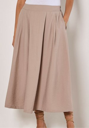Jupe midi beige avec un large pli devant, taille haute et poches latérales, portée avec des sandales à lacets couleur cuir clair et un haut blanc.