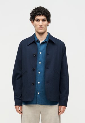 Homme aux cheveux bouclés portant une veste bleu marine sur une chemise bleue à boutons et un pantalon clair, debout devant un fond blanc uni.