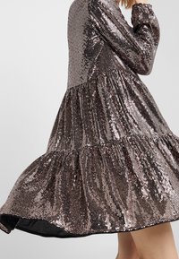 Robe brillante et pailletée dans des nuances de bronze et de marron foncé. Caractéristiques d'une jupe à niveaux et de longues manches avec des poignets froncés. Texture lisse.