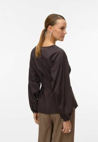 Dunkelbraune Bluse mit langen, puffigen Ärmeln und taillierter Passform. Das Material wirkt glatt mit einem subtilen Glanz. Auffällig sind die klaren Linien.