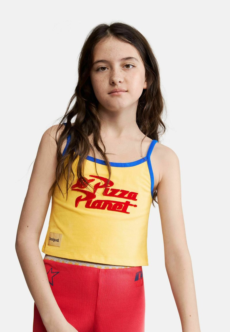 Fille aux longs cheveux foncés portant un débardeur jaune « Pizza Planet » avec un liseré bleu et un pantalon rouge, sur un fond clair uni.