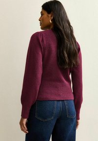Maglione a maglia color bordeaux con maniche lunghe, orlo a coste e finitura testurizzata, abbinato a jeans in denim scuro. La vista posteriore mostra una leggera sagomatura.