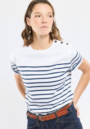 Femme portant un chemisier à manches courtes rayé blanc et bleu marine avec des boutons sur l'épaule, des boucles d'oreilles créoles dorées, une ceinture marron et un jean foncé.