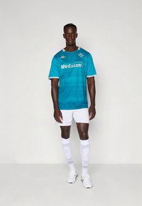 Camisola de futebol turquesa com design em gradiente, detalhes brancos e logótipo. Combinada com calções brancos, meias brancas até ao joelho e ténis.