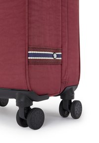 Red fabric suitcase with a textured surface, accented by a brown stripe and blue detailing. Features four black wheels for mobility.
