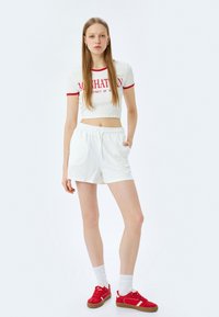 Witte katoenen shorts met een elastische tailleband en zakken, gecombineerd met een wit crop top met korte mouwen en een geribbelde structuur, voorzien van rode letters. Rode sneakers.
