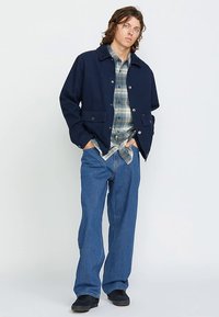 Marineblaue Jacke mit Kragen, Fronttaschen und Knopfverschluss, kombiniert mit einem karierten Hemd und blauen Jeans. Flache Schuhe vervollständigen den Look.