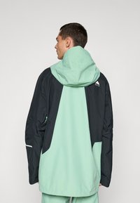 Lett jakke med mintgrønn og svart fargeskjema, med hette, glidelåser og reflekterende detaljer. Glatt, funksjonell design.