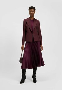 Burgundowy dwurzędowy żakiet nałożony na dopasowaną plisowaną sukienkę. Teksturowana tkanina, spódnica do kolan, uzupełniona czarnymi kozami za kolano.