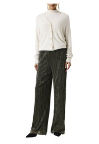 Vrouw in een witte fuzzy knoopcardigan, metalen geplooide wijde broek, zwarte puntige schoenen en lange gouden oorbellen.