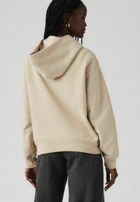 Sudadera beige con un gran bolsillo trasero y capucha ajustable, combinada con jeans gris oscuro. Textura suave y corte relajado.