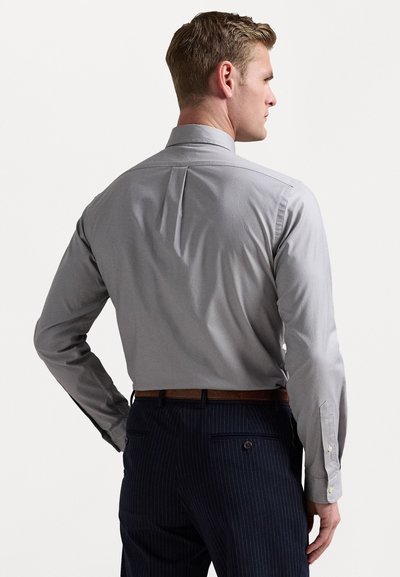 Maglietta grigia chiara a maniche lunghe con piega sul retro, abbinata a pantaloni scuri a righe. La maglietta presenta polsini con bottoni e colletto standard.