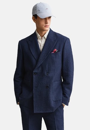 Man draagt een marineblauw gestructureerd dubbelbreasted pak met een rood pochet, een beige overhemd en een lichtblauwe gestreepte baseballpet, handen in de zakken.