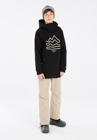 Schwarze mittellange Jacke mit grafischen Wellen in Beige, kombiniert mit beigen Hosen und schwarzen Stiefeln, ausgestattet mit einer Kapuze und elastischen Bündchen.