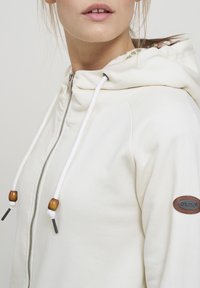 Sudadera crema con cremallera frontal, capucha y costuras decorativas; cuenta con cuentas de madera en los cordones y una etiqueta con el logotipo.