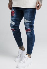 Vaqueros de mezclilla con un lavado oscuro, detalles desgastados, parches rasgados y acentos florales rojos. Combinados con zapatillas blancas con diseño de cordones.