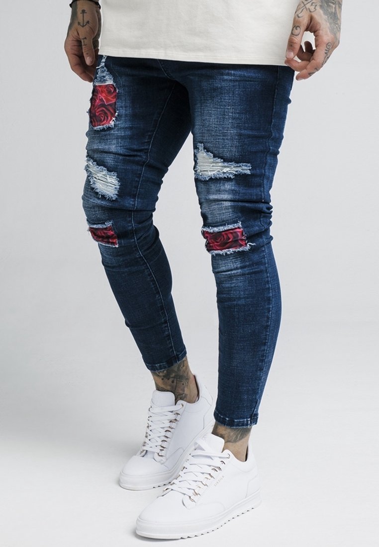 Vaqueros de mezclilla con un lavado oscuro, detalles desgastados, parches rasgados y acentos florales rojos. Combinados con zapatillas blancas con diseño de cordones.