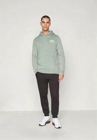 Um hoodie verde claro com um bolso tipo canguru e logotipo, combinado com calças de moletom pretas e ténis brancos atléticos com detalhes em vermelho.