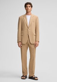 Beige gestreifter Anzug mit einem einreihigen Blazer, passenden Hosen und einem weißen T-Shirt darunter, ergänzt durch schwarze Sandalen.