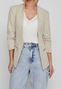 Blazer beige a medida con mangas largas, frente abierto y detalles con cremallera. Lleva puesto sobre una camiseta blanca y vaqueros de mezclilla azul de tiro alto.