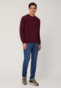 Maglione a maglia bordeaux con maniche lunghe e scollo rotondo; abbinato a jeans in denim blu e sneakers multicolori con dettagli testurizzati.