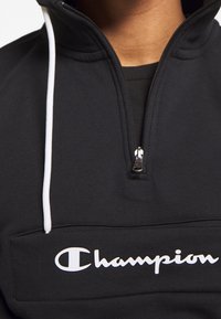 Felpa nera in pile con mezza zip e lacci bianchi. Presenta una tasca frontale e un prominente logo bianco "Champion". Tessuto liscio.