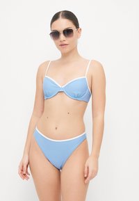 Lys blå bikiniset med innlegg i undertrådstoppen og høyt liv-bukser, begge trimmet med hvite detaljer. Modell med solbriller.