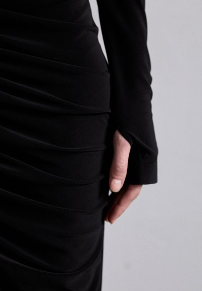 Black dress with long sleeves, featuring gathered fabric for texture. Close-up shows hand resting on the side, emphasizing the draped design.
