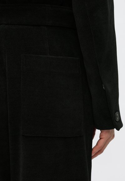 Fekete kordbársony ruhadarab nagy foltzsebbel és gombos mandzsettával. A textúra bordázott, puha, tapintható felületet biztosít.