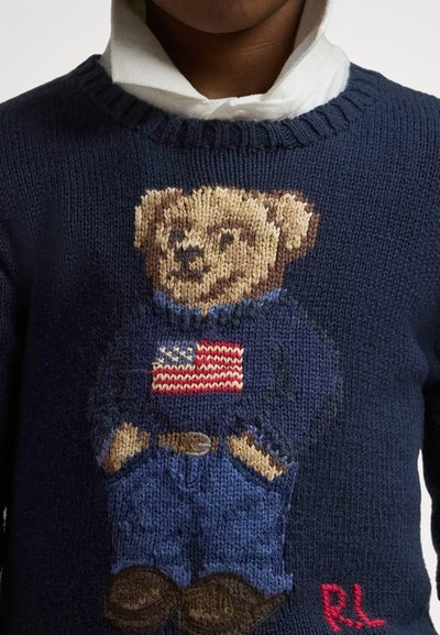 Strikket navygenser med bjørn som har på seg genser med amerikansk flagg og jeans, brukt over en hvit skjorte med synlig krage.