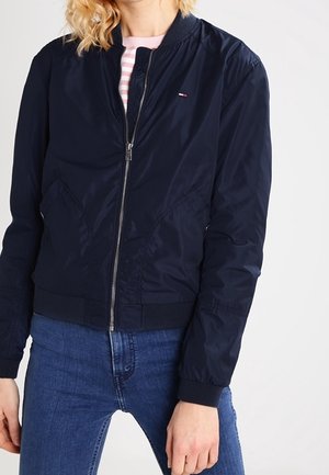 Kvinde iført en marineblå bomberjakke med frontlynlås, lommer og et lille logo på brystet, kombineret med blå jeans og en stribet skjorte.