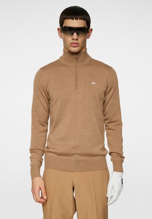 Maglione marrone a mezza zip realizzato in tessuto morbido, con silhouette aderente e polsini a coste. Dettaglio del logo bianco sul petto, abbinato a pantaloni beige e un guanto bianco.